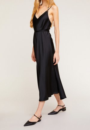 Kobieta idąca w czarnej, bezrękawnikowej sukience midi na cienkich ramiączkach i czarnych butach typu slingback na niskim obcasie z ostro zakończonym noskiem.