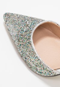 Glittrig silver högklackad sko med spetsig tå och öppen baksida, som visar en glänsande yta och ett beige inre.