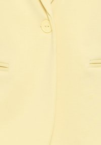 Un blazer jaune pâle avec une seule boutonnière jaune et deux poches fendues horizontales à l'avant.