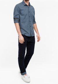 Hombre con una camisa azul de patrón enrollada, jeans oscuros con dobladillo y zapatillas blancas, de pie contra un fondo blanco liso.