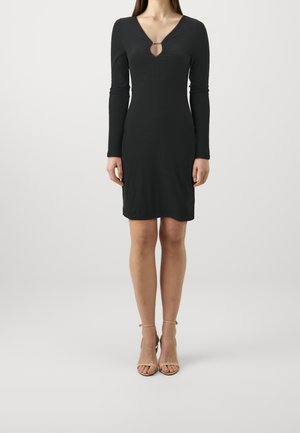 Woman wearing a fitted black long-sleeve dress with a keyhole neckline and nude ankle-strap heels, standing against a plain background.