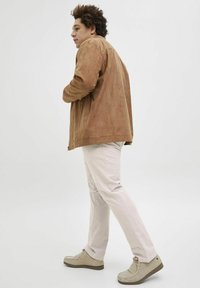 Veste en daim marron, pantalon beige et chaussures gris clair. La veste arbore une coupe décontractée et un design minimaliste, offrant une texture lisse.