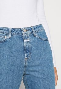 Jeans de mezclilla en azul claro con un diseño de cintura alta. Cuenta con un cierre de botón metálico, costuras de cobre y bolsillos frontales.