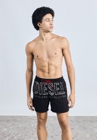 Mustad ujumispüksid, millel on punane nöör, suur valge "DIESEL" logo ja tekst "FOR SUCCESSFUL LIVING". Sile kangas, keskmise reie pikkus.