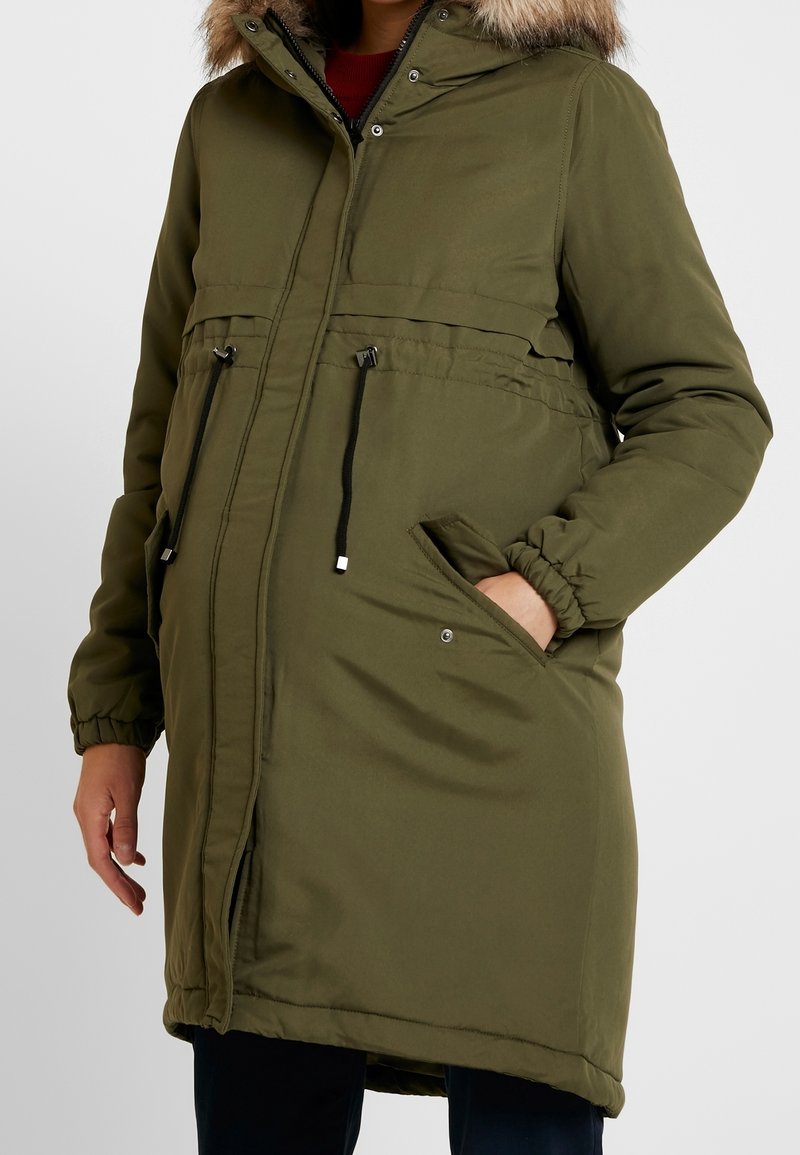 Olivgrön vadderad vinterjacka med huva prydd med päls, svarta dragskor i midjan, frontdragkedja och sidofickor med tryckknappar.
