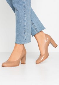 Escarpins en cuir verni beige avec un bout arrondi et un talon carré. Portés avec un jean en denim bleu clair coupé qui présente des ourlets effilochés.