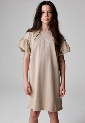 Beige bomullsklänning med kort puffärmar, med en cirkulär logotyp i ljusblått som har texten "SAINT LYS LONDON" på framsidan.