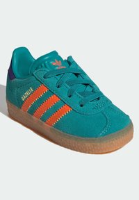 Teal mockask sneaker med orange och gula randiga detaljer, med en gummisula och en vadderad krage. Modellenamn "Gazelle" är synligt.