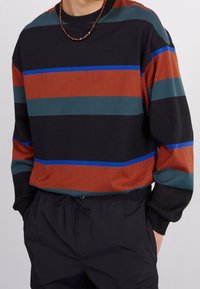 Mikina s dlouhými rukávy a proužky v černé, rezavé, zelené a modré barvě. Měkká tkanina, volný střih, s žebrovaným výstřihem a manžetami.
