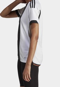 White athletic jersey featuring black accents, three black stripes on the shoulders, and a textured fabric. Black shorts are visible below.