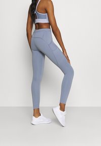 Leggings bleu clair avec une taille haute, texture lisse, poches latérales et coutures contrastées, portés avec des chaussures de sport blanches.