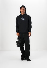 Felpa nera con logo, pantaloni neri larghi e sneakers nere. L'uomo tiene una piccola borsa a tracolla nera con tracolla regolabile.