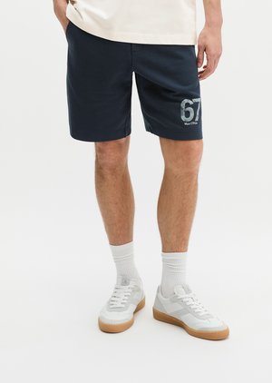 Mężczyzna w granatowych szortach z nadrukiem „67 Marc O'Polo”, białych skarpetkach i białych tenisówkach z gumową podeszwą, stojący na jednolitym białym tle.