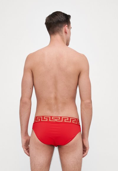 Rote Schwimmslips mit einem goldenen griechischen Schlüssel-Muster am Bund, mit glatter Textur und enganliegendem Design. Rückansicht.