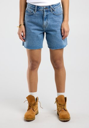 Persoon die blauw denim shorts van middellange lengte draagt, witte sokken, bruine werklaarzen en een gouden armband, staand tegen een effen achtergrond.