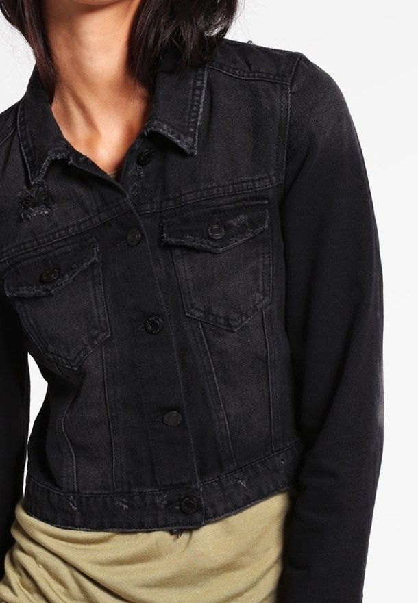 Chaqueta de mezclilla negra con dos bolsillos en el pecho, cierre frontal con botones, bordes deshilachados y diseño corto; combinada con una camiseta de color claro debajo.