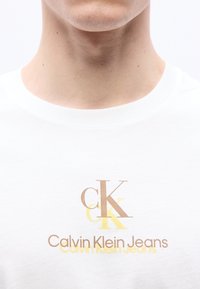 Κοντινό πλάνο του λαιμού και του στήθους ενός ατόμου που φοράει άσπρο πουκάμισο με το λογότυπο "CK" και "Calvin Klein Jeans" σε καφέ και κίτρινο χρώμα.