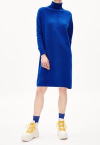 Blå stickad klänning med hög krage, långa ärmar och rak skärning; bärs med vita hög- topp sneakers och blå strumpor med gula snören.