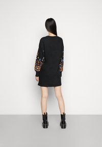 Robe noire avec des manches longues à motifs ornées de broderies florales colorées. Le mannequin porte des bottines noires ; texture de tissu lisse.