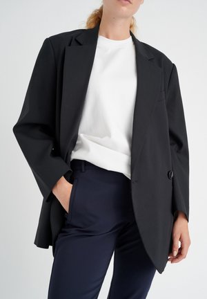 Personne portant un blazer noir oversize, un t-shirt blanc et un pantalon bleu marine, avec une main dans la poche sur un fond clair uni.