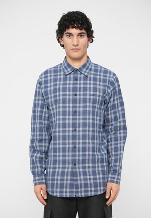 Młody mężczyzna z kręconymi włosami, ubrany w niebieską i szaro-kratkowaną koszulę z guzikami, stoi na tle jednolitego białego tła.