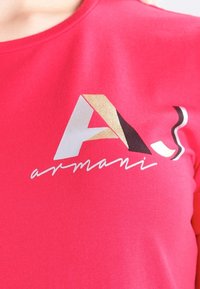 Różowa bawełniana koszulka z nadrukiem w wielokolorowym logo, zawierającym tekst "A" oraz "armani" w białych, czarnych, złotych i srebrnych akcentach.