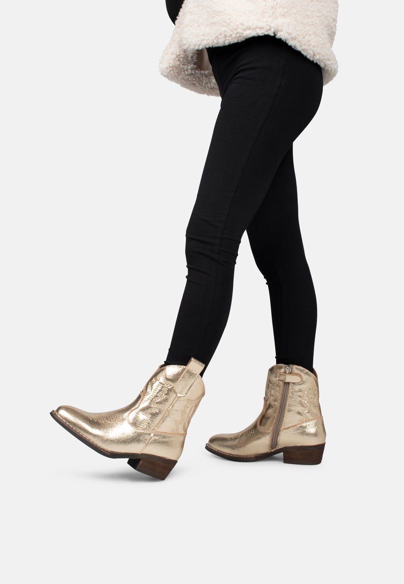 Botas tobilleras metálicas doradas con un acabado texturizado, cremallera lateral y un bajo tacón de madera, combinadas con leggings negros. Enfoque en los detalles del calzado.
