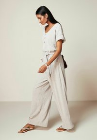 Jumpsuit beige chiaro e strutturato, con scollatura a V, maniche corte, bottoni frontali, vita cinturata e gambe larghe, abbinato a sandali piatti semplici.