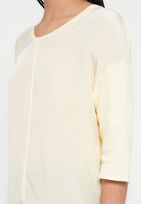 Top galben deschis cu mâneci lungi, gât în V și un luciu subtil. Țesătura moale are o cusătură centrală și manșete scurte rulate.