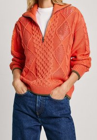 Suéter de punto naranja con cuello alto, que presenta patrones de rombos y texturas de punto cableado. Bajo y puños acanalados con un acento verde en contraste.