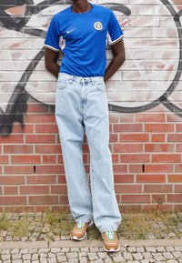 Camiseta azul de Nike del Chelsea con acentos en blanco y dorado, jeans holgados color azul claro y zapatillas color marrón con detalles en verde, todo frente a una pared de ladrillo.