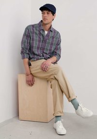 Camisa a cuadros en púrpura y verde, chinos beige, zapatillas blancas, calcetines azul claro, gorra azul marino, sentado sobre un bloque de madera.