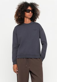 Kvinne med krøllete hår iført mørke oversized solbriller, en kullgrå sweatshirt og brune joggebukser, som står mot en ensfarget bakgrunn.