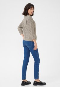 Helles graues Strickpullover mit Rautenmuster, Dreiviertelärmel, kombiniert mit blauen Skinny-Jeans und schwarzen Slippern, von hinten betrachtet.