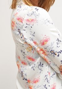 Bloemenblouse van lichte stof, met roze en gele bloemen op een witte achtergrond, met blauwe accenten en een gladde textuur.