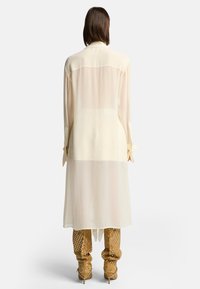 Camicia lunga di un leggero beige chiaro con colletto e maniche con bottoni, che si appoggia fino a metà coscia, abbinata a stivali tan a fantasia.