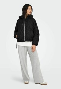 Gesteppte schwarze Jacke mit Kapuze, weißem Reißverschluss und Kordelzug. Kombiniert mit hellgrauen, locker sitzenden Hosen und beigen Schuhen.