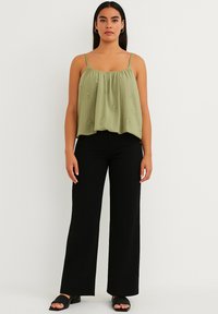 Top senza maniche di colore verde oliva con scollatura arricciata e dettagli ricamati, abbinato a pantaloni neri a vita alta e sandali neri.