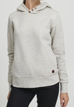 Femme portant un sweat à capuche gris clair à pois avec trois boutons en bois près du col et un pantalon noir.