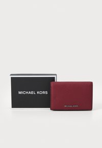 Portafoglio in pelle rossa con finitura testurizzata, caratterizzato da un logo "MICHAEL KORS" in argento. Presentato accanto a una scatola nera del marchio con scritte bianche.