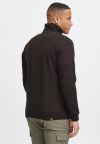 Mørkebrun sweatshirt med høy krage, ribbede mansjetter og kant, glatt stoff, enkelt design og et lite logo-merke på baksiden.