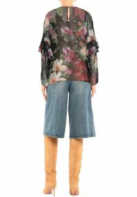 Person, der eine transparente schwarze Bluse mit Blumenmuster, knielange Jeansshorts und beiger, hochhackiger Wildlederstiefel trägt, mit dem Rücken zum Betrachter.