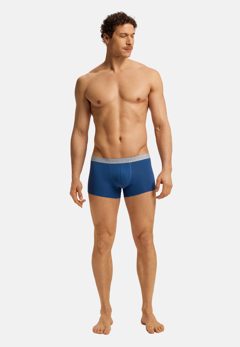 Homme musclé aux cheveux bruns bouclés portant un boxer bleu avec une ceinture grise, debout pieds nus sur un fond blanc.