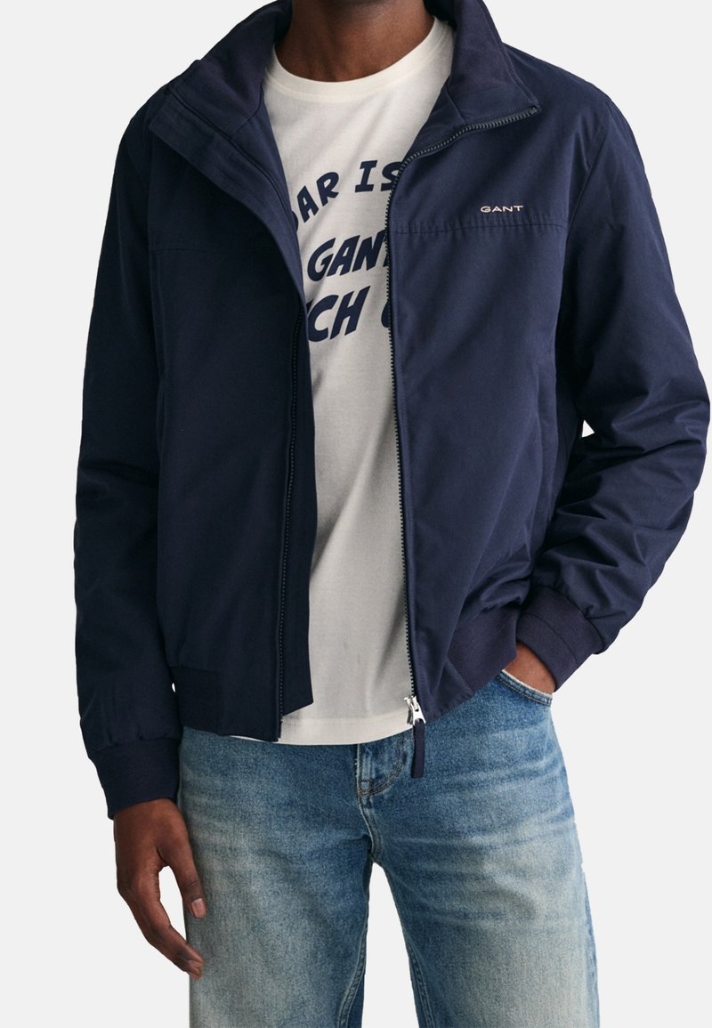 Giacca bomber blu navy con zip frontale, polsini a coste e dettaglio del logo. Indossata sopra una t-shirt grafica grigia chiara e jeans in denim.