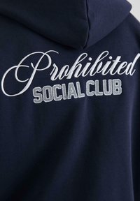 Marineblå hættejakke med hvid broderet tekst "Prohibited" i kursiv og "SOCIAL CLUB" i blokbogstaver med en tekstureret finish.