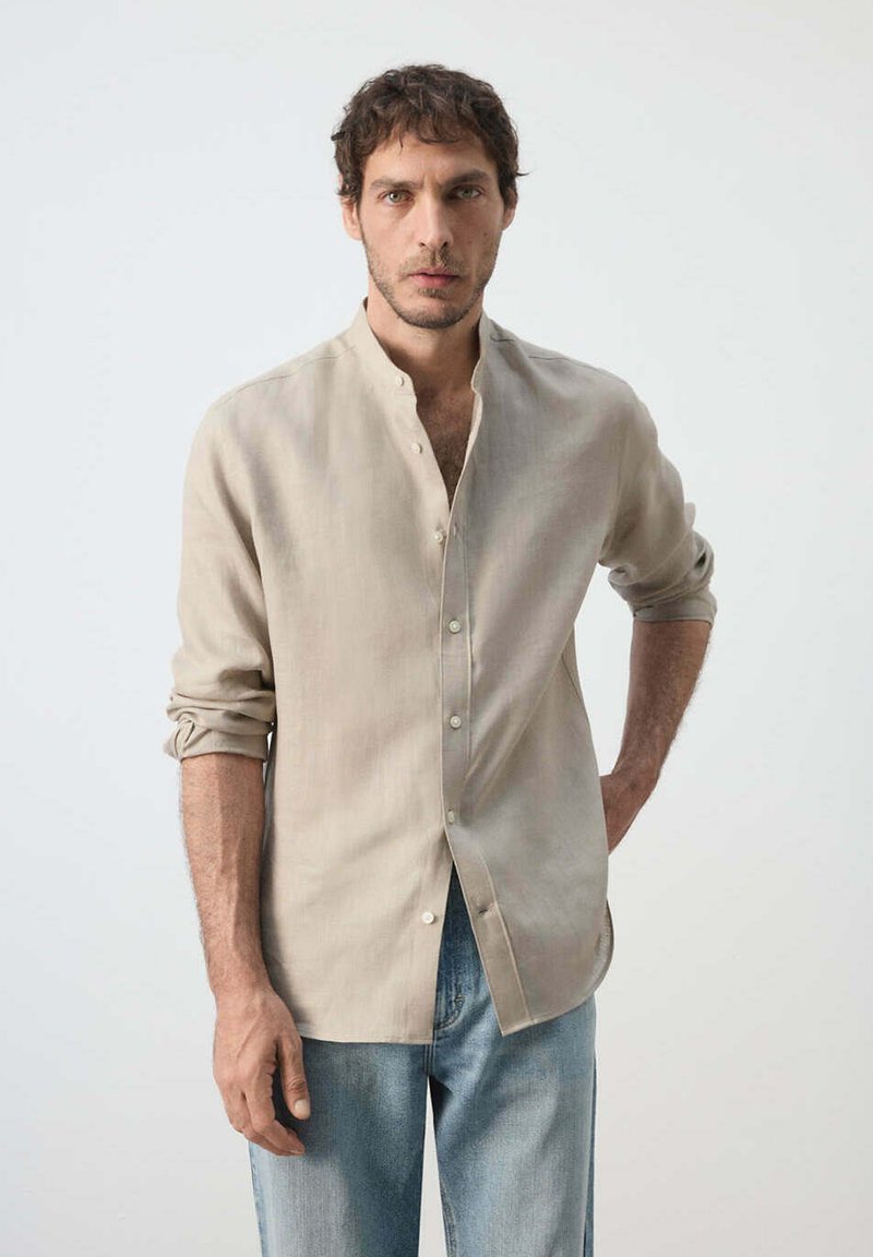 Homme aux cheveux foncés portant une chemise beige à boutons avec manches retroussées et jean bleu, debout devant un fond clair uni.