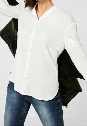 Kvinde iført en enkel hvid skjorte med knapper og blå jeans, der holder en sort jakke bag ryggen mod en ensfarvet baggrund.