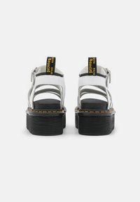 Hvite platå sandaler med svarte teksturerte såler, åpent design og flere stropper. Har gul tekst på heleklaffen.