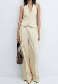 Gilet beige su misura con scollo a halter e tre bottoni, abbinato a pantaloni a gamba larga. Accessoriato con una borsa a spalla marrone.