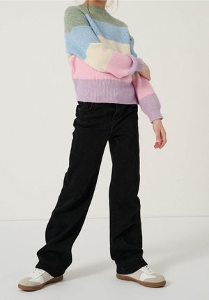 Personne portant un pull rayé pastel, un pantalon noir ample et des baskets blanches, debout devant un fond uni.